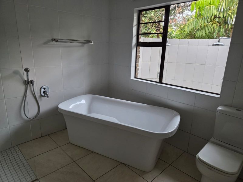 A clean, modern bathroom featuring a freestanding bathtub, a toilet, and a shower area with tiled finishes and a window overlooking the garden.