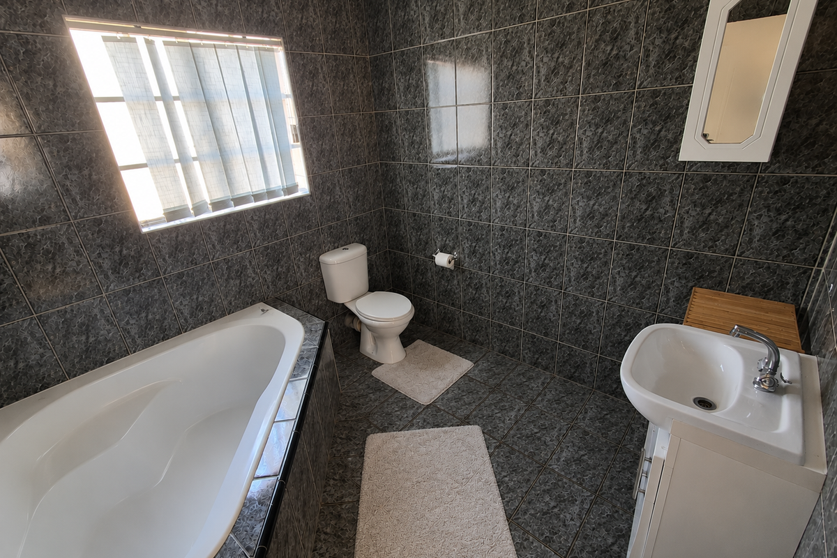 A functional bathroom featuring dark marble-effect wall and floor tiles, a built-in corner bathtub, a white vanity basin, and a wall-mounted mirror.