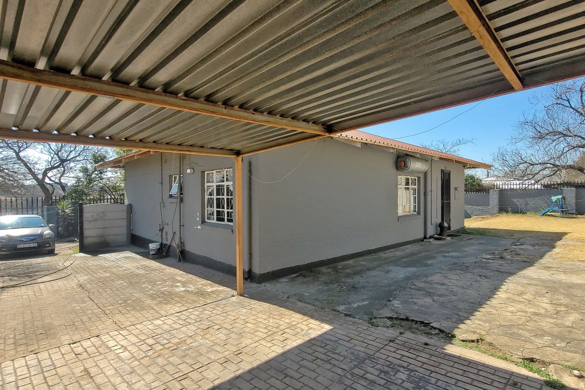 Rear exterior view of the house featuring a large corrugated metal carport, paved driveway, and external geyser unit.