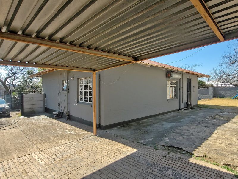 Rear exterior view of the house featuring a large corrugated metal carport, paved driveway, and external geyser unit.