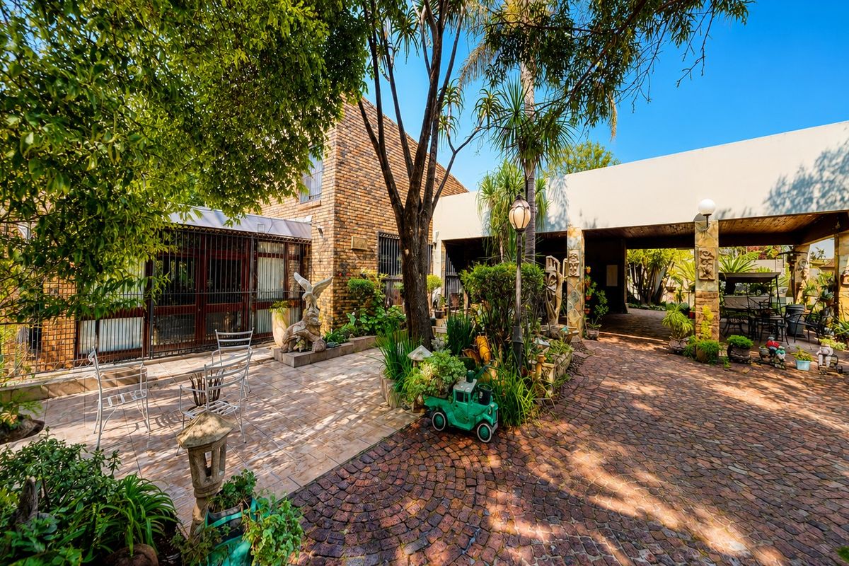 A spacious rear courtyard featuring brick paving, a tiled patio area, and a covered carport, surrounded by established garden landscaping.