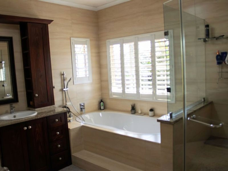 A well-appointed bathroom featuring a built-in vanity with stone countertop, a deep soaking bathtub, and a separate glass-enclosed shower with plantation shutters.