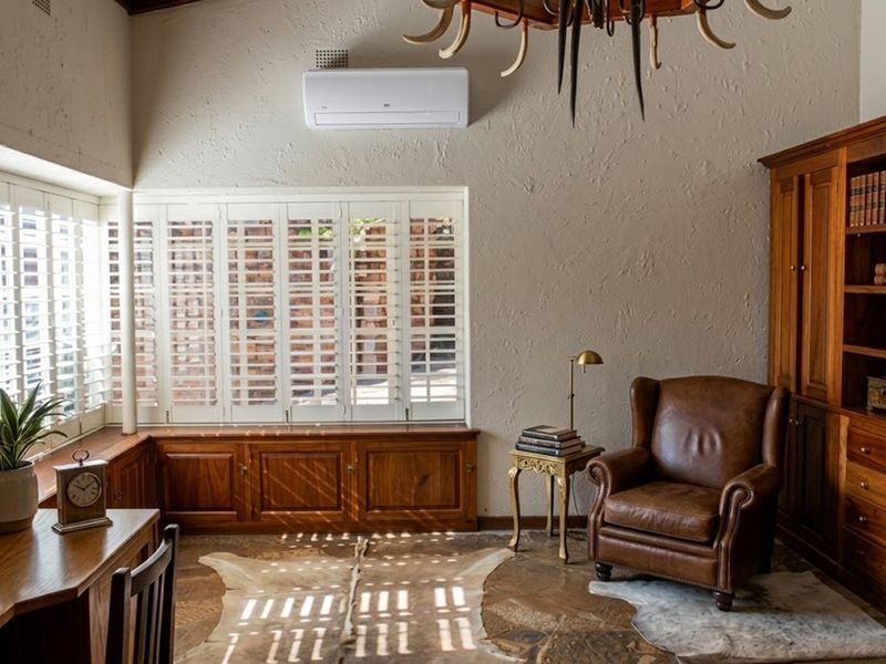 A well-appointed home office featuring custom built-in wooden cabinetry, integrated shelving, stone flooring, and plantation-style window shutters for adjustable natural light.
