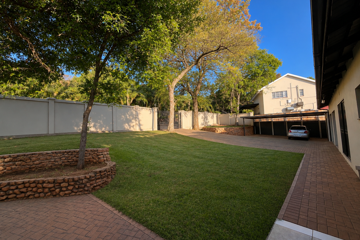 Spacious rear garden featuring a well-maintained lawn, paved driveway, stone retaining wall, and a covered carport area.