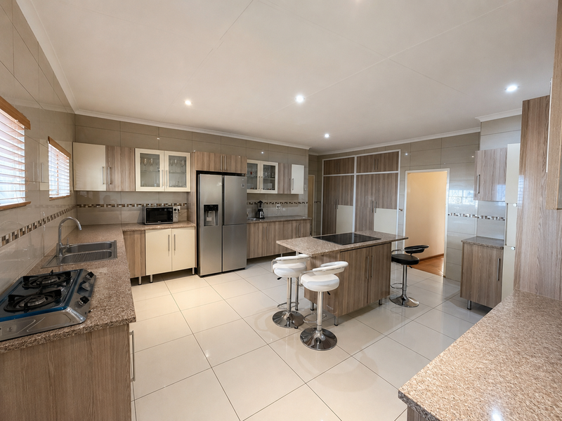 Spacious modern kitchen featuring granite countertops, ample built-in cabinetry, a central island with a gas hob, and neutral tiled flooring throughout.