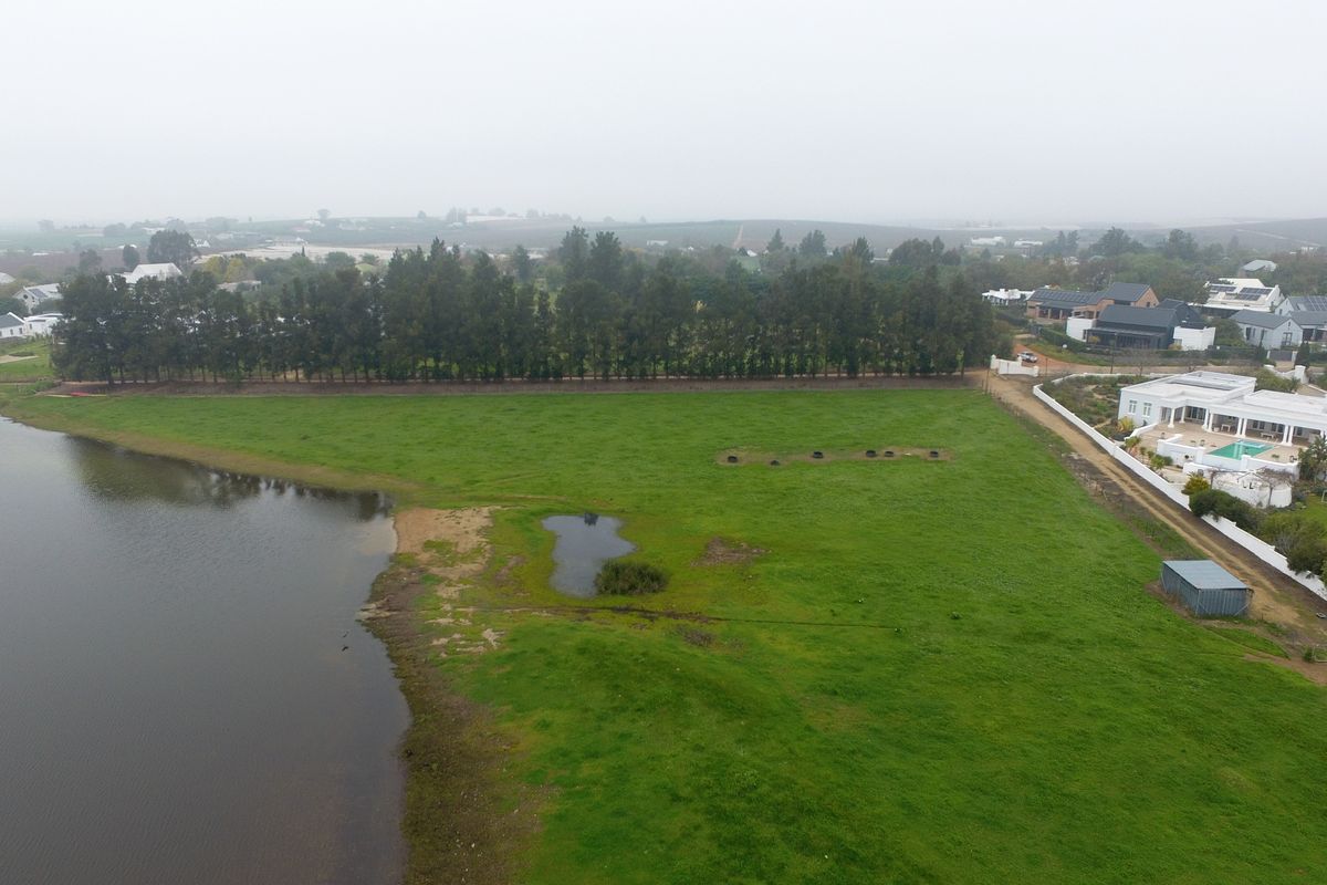 An aerial view of a vacant residential plot featuring direct lake frontage, established perimeter trees, and surrounding residential development.