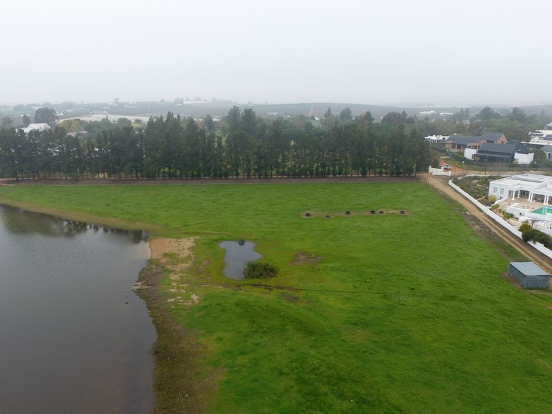 An aerial view of a vacant residential plot featuring direct lake frontage, established perimeter trees, and surrounding residential development.