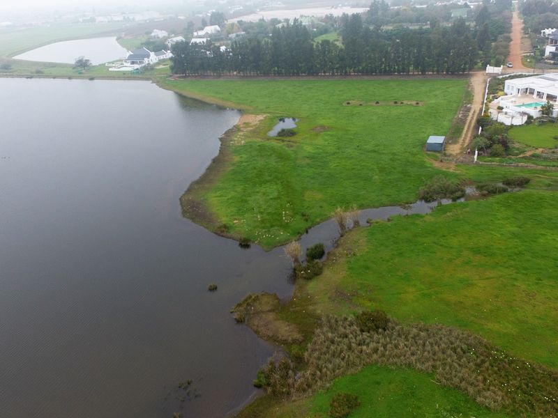 An aerial view of a vacant residential land plot featuring expansive green fields, a natural lake, and proximity to established residential properties.