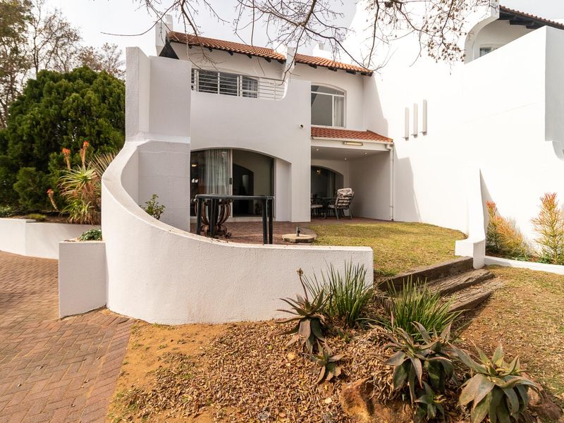Rear view of the property featuring a covered patio, paved driveway, and landscaped garden area with established trees and retaining walls.