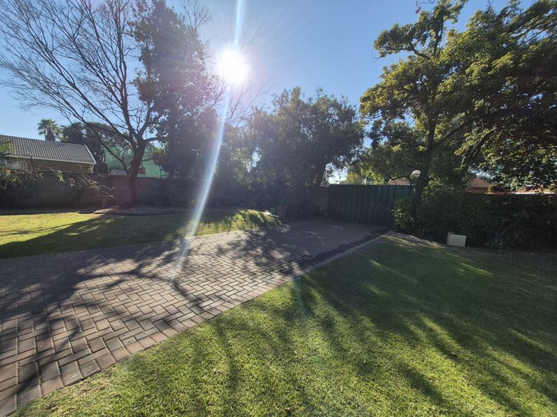 A spacious garden area featuring a paved driveway, established trees, and secure perimeter walling with a metal gate.