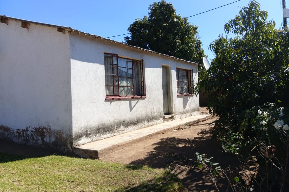 Rear exterior view of a single-story house showing weathered white plaster walls, metal window frames, and a concrete pathway.