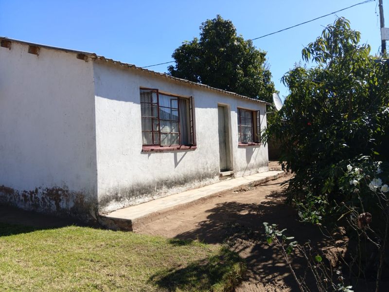 Rear exterior view of a single-story house showing weathered white plaster walls, metal window frames, and a concrete pathway.