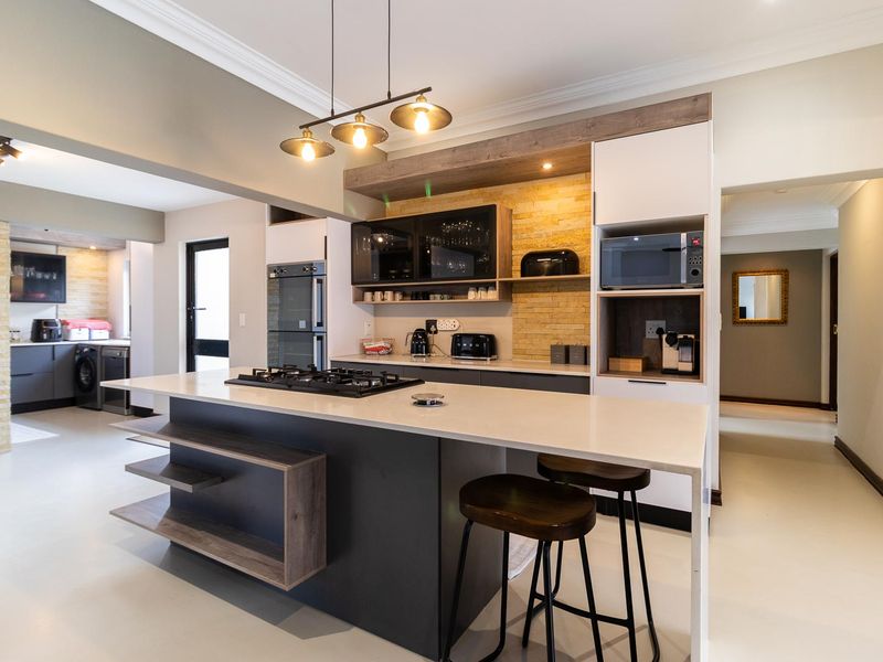 Modern open-plan kitchen featuring a large island with gas cooktop, stone countertops, stacked stone accents, and integrated appliances, flowing into a functional scullery area.