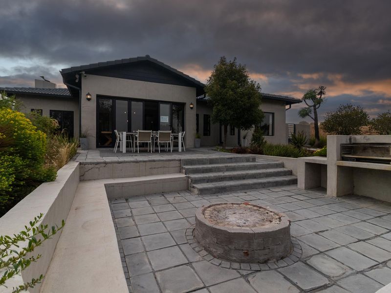 Rear exterior view featuring a paved patio, built-in braai, stone fire pit, and concrete steps leading to the main house entrance.