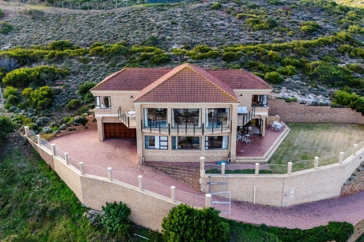 Aerial view of a multi-level brick residence featuring a tiled roof, expansive paved driveway, perimeter retaining walls, and a balcony overlooking the surrounding landscape.