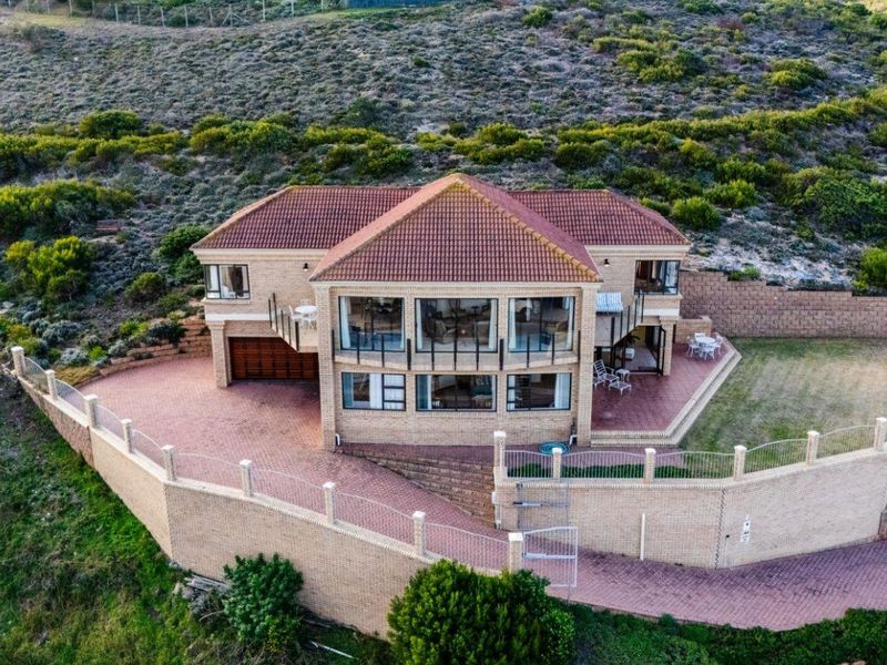 Aerial view of a multi-level brick residence featuring a tiled roof, expansive paved driveway, perimeter retaining walls, and a balcony overlooking the surrounding landscape.