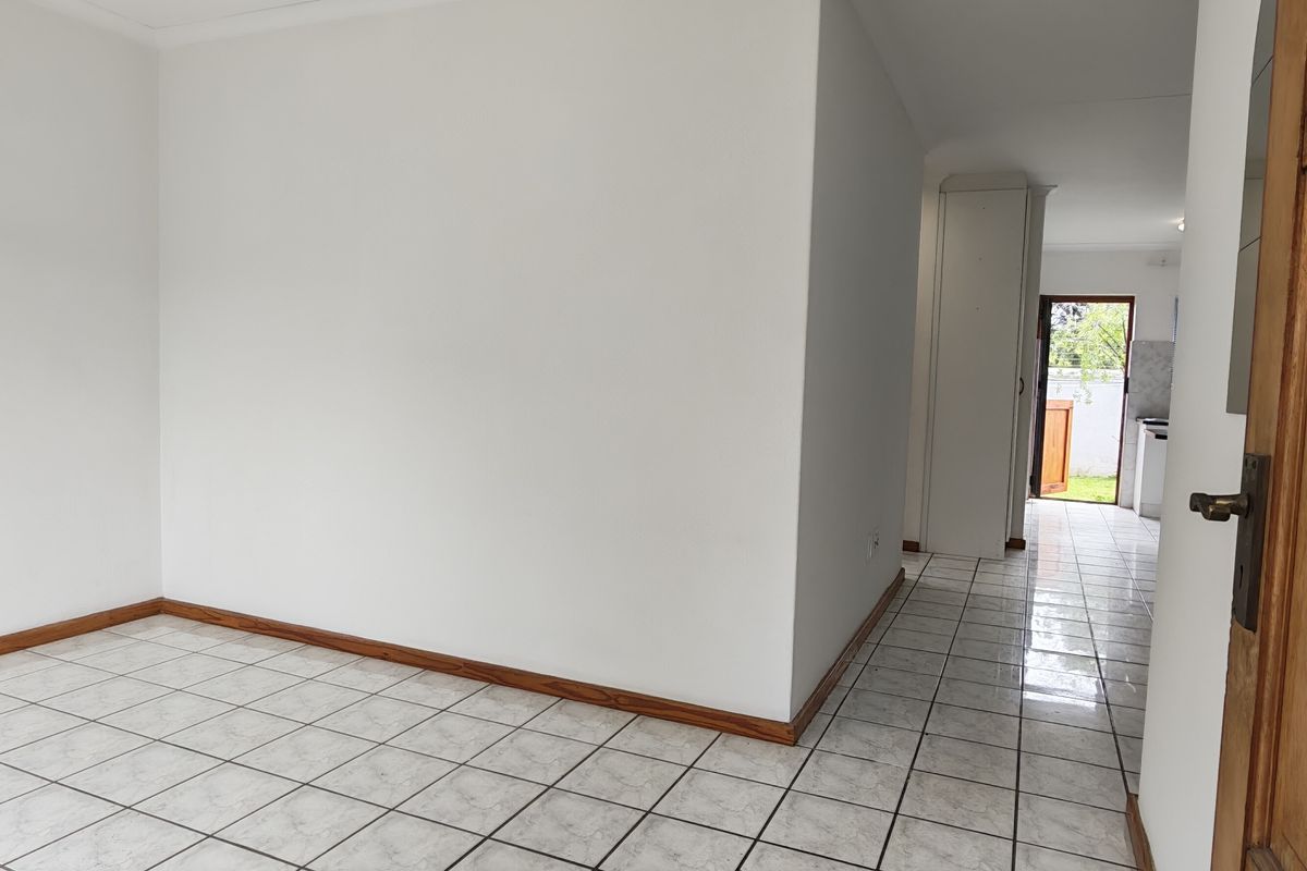 An empty, neutral-toned living area featuring ceramic tile flooring and timber skirting, with a clear view through to the rear entrance.