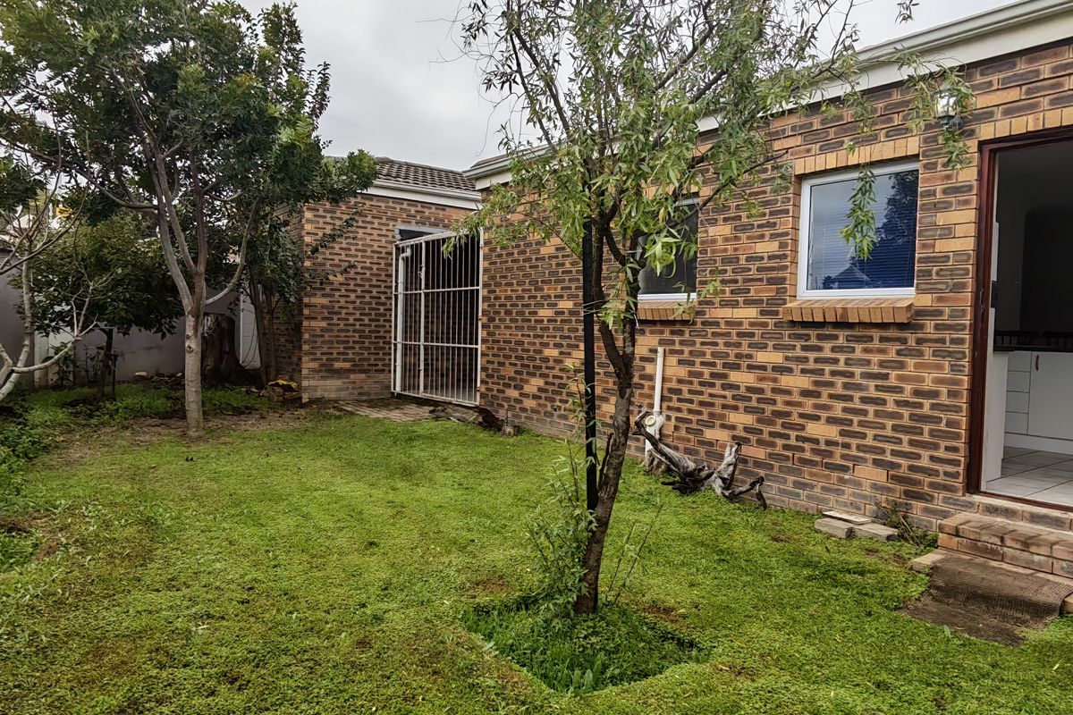 A rear garden view featuring a face brick exterior, a timber door entrance, and a lawn area with established trees and boundary walling.