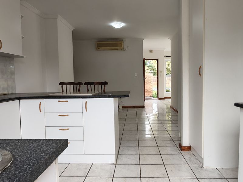 Open-plan kitchen featuring white cabinetry, dark laminate countertops, tiled flooring, and a wall-mounted air conditioning unit, leading into an adjacent living area.