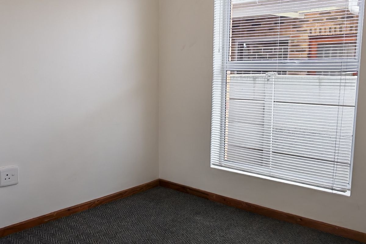 A clean, empty bedroom featuring neutral walls, dark patterned carpeting, a ceiling fan, and venetian blinds on the window.