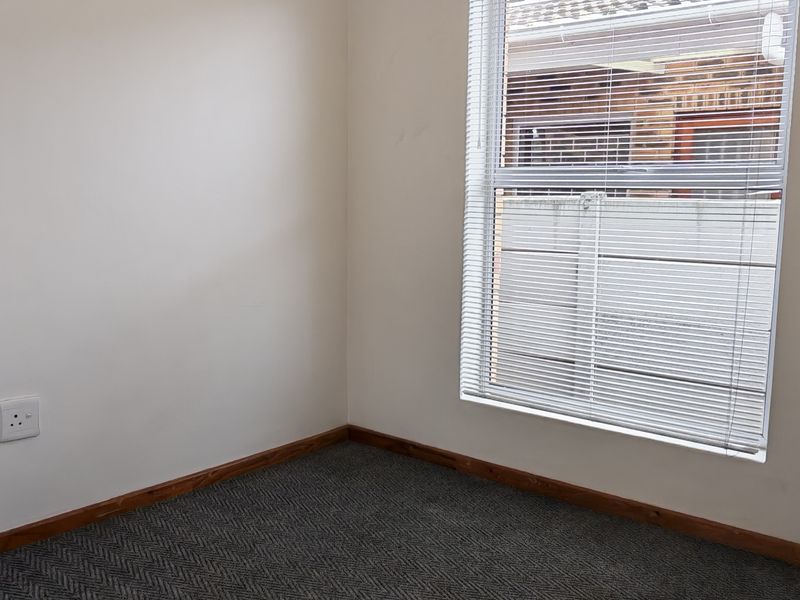 A clean, empty bedroom featuring neutral walls, dark patterned carpeting, a ceiling fan, and venetian blinds on the window.
