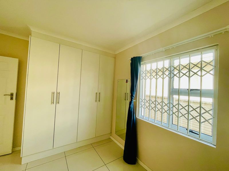 A clean, empty bedroom featuring white built-in cupboards, tiled flooring, and a window fitted with a security gate.