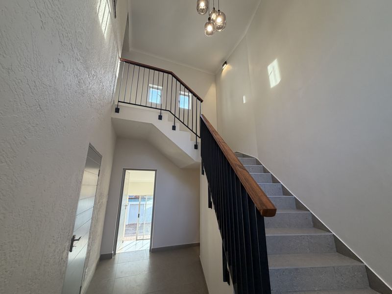 A bright, modern double-volume entrance hallway featuring a tiled staircase with a sleek metal balustrade and wooden handrail, leading to an upper level.