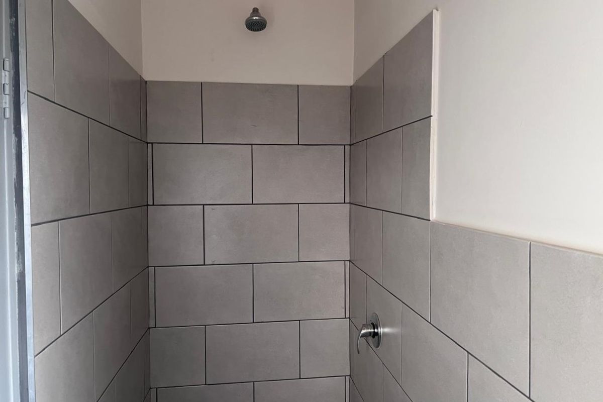 A shower enclosure featuring grey subway-style wall tiles, a fixed shower head, and a chrome mixer tap, currently showing incomplete tiling on the side wall.