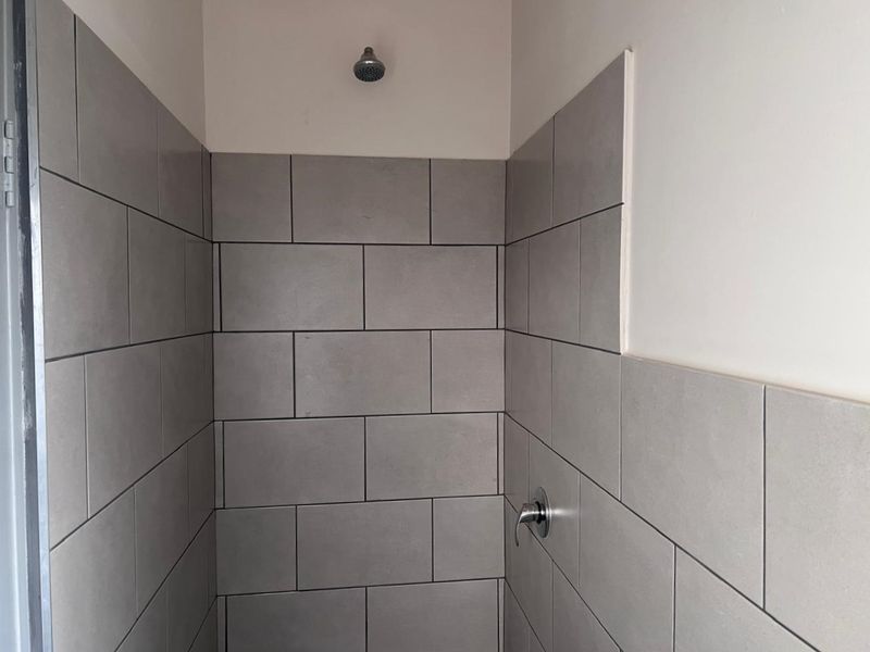 A shower enclosure featuring grey subway-style wall tiles, a fixed shower head, and a chrome mixer tap, currently showing incomplete tiling on the side wall.