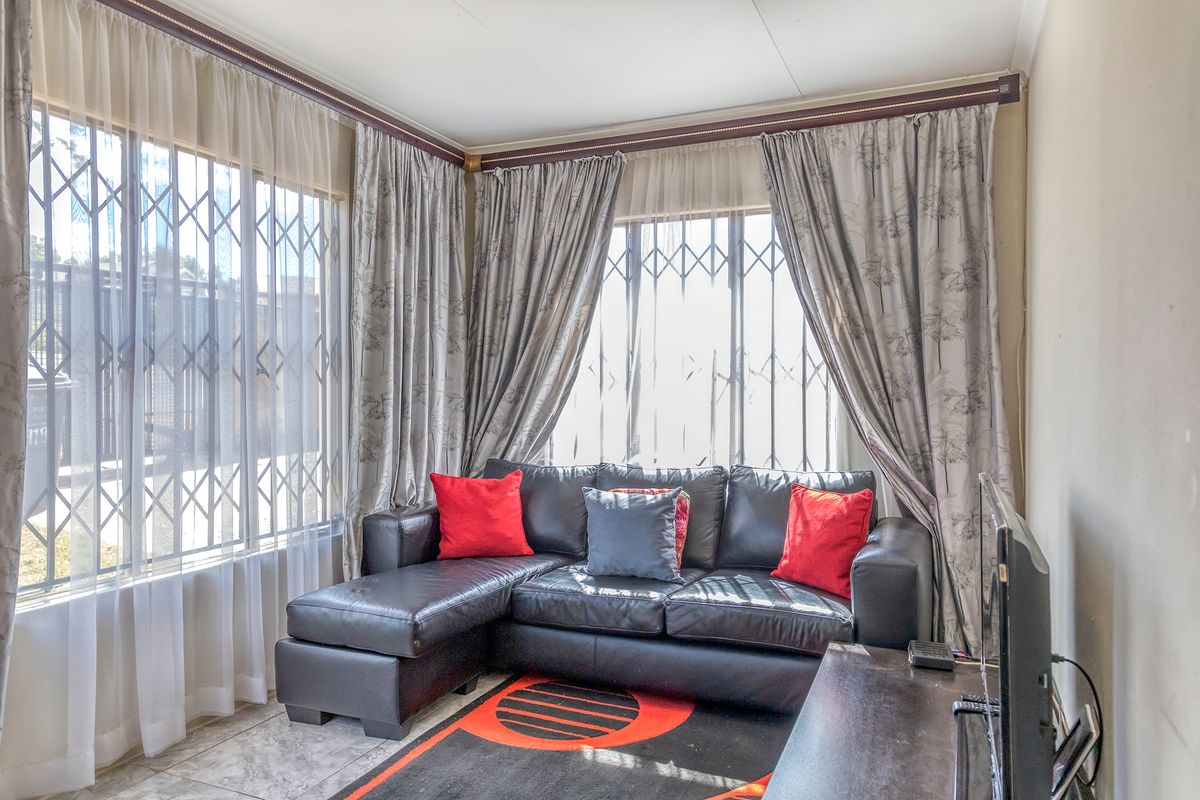A bright living room featuring tiled flooring and large windows fitted with security gates and curtain rails.