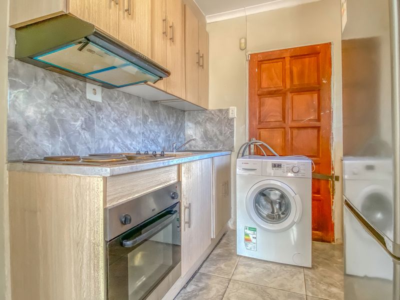 Compact kitchen featuring light wood cabinetry, marble-effect splashback, built-in oven and hob, and tiled flooring with direct access to an exterior door.