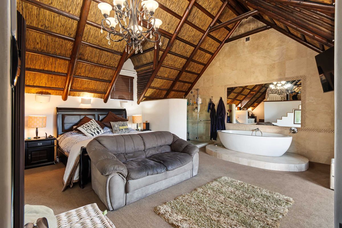 Spacious bedroom featuring a high vaulted thatched ceiling with exposed rafters, integrated ensuite bathroom with a freestanding tub, and stone wall finishes.