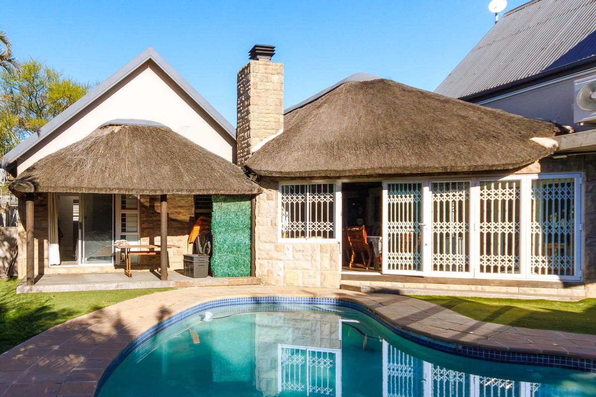 Rear exterior view featuring a swimming pool, stone-clad walls, and traditional thatched roofing with security gates leading into the home.