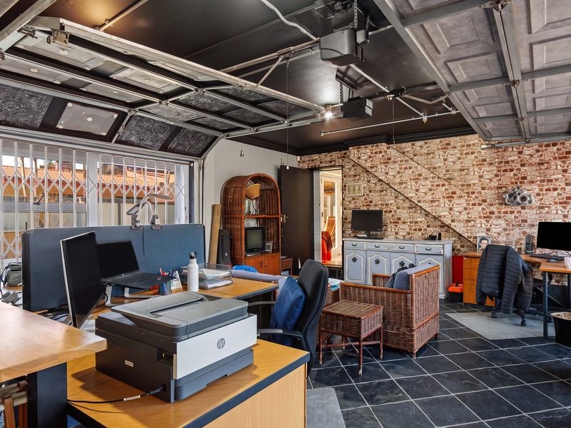 A converted garage space featuring exposed brick walls, black tiled flooring, and an industrial-style ceiling, currently utilized as a multi-purpose workspace.