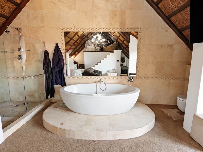 Spacious bathroom featuring a central freestanding bathtub on a raised platform, glass-enclosed shower, and natural stone wall tiling under a vaulted thatch ceiling.