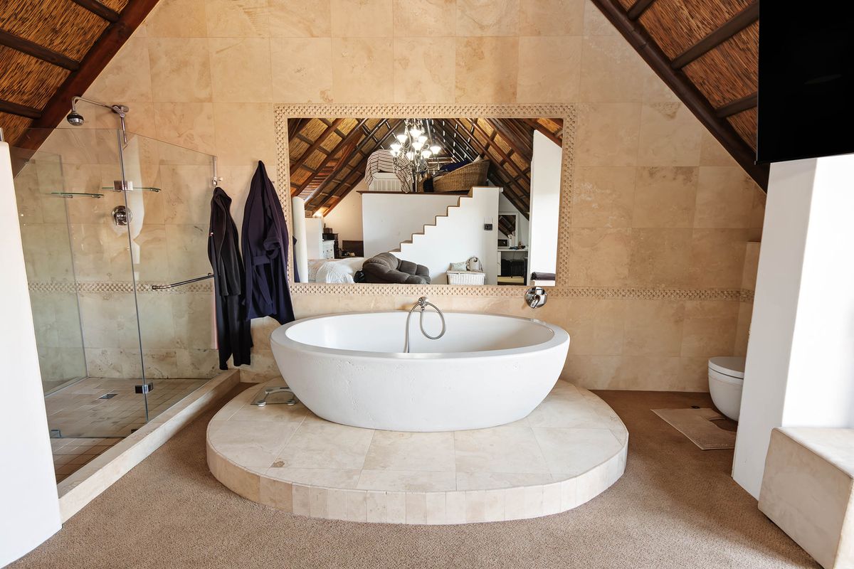 Spacious bathroom featuring a central freestanding bathtub on a raised platform, glass-enclosed shower, and natural stone wall tiling under a vaulted thatch ceiling.