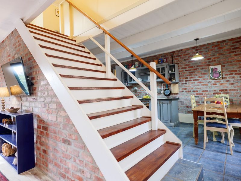 Open-plan living and dining area featuring exposed brick walls, slate flooring, timber ceiling beams, and a central staircase leading to the upper level.