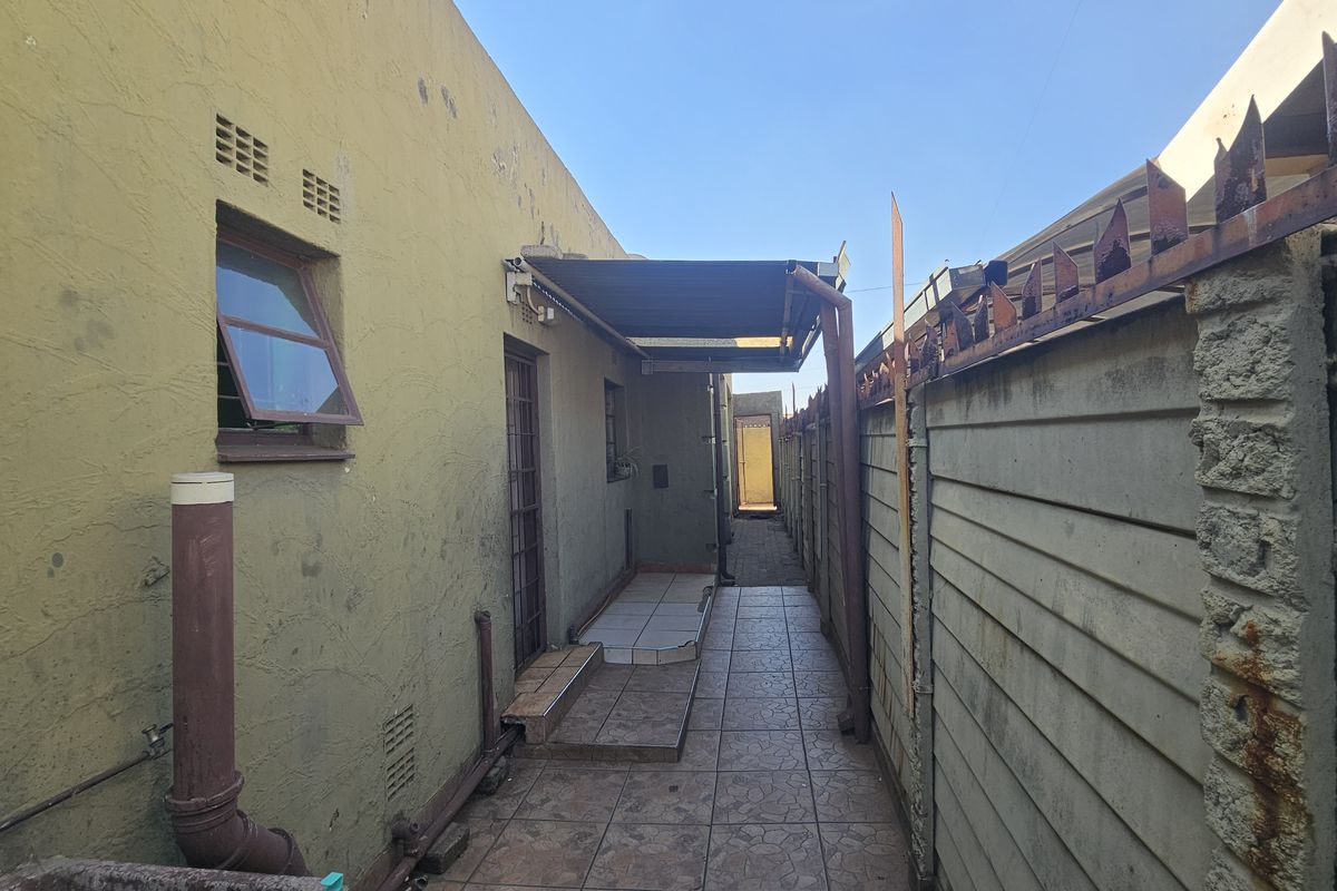 A narrow rear exterior walkway featuring tiled flooring, a covered section, and a precast concrete boundary wall with security spikes.