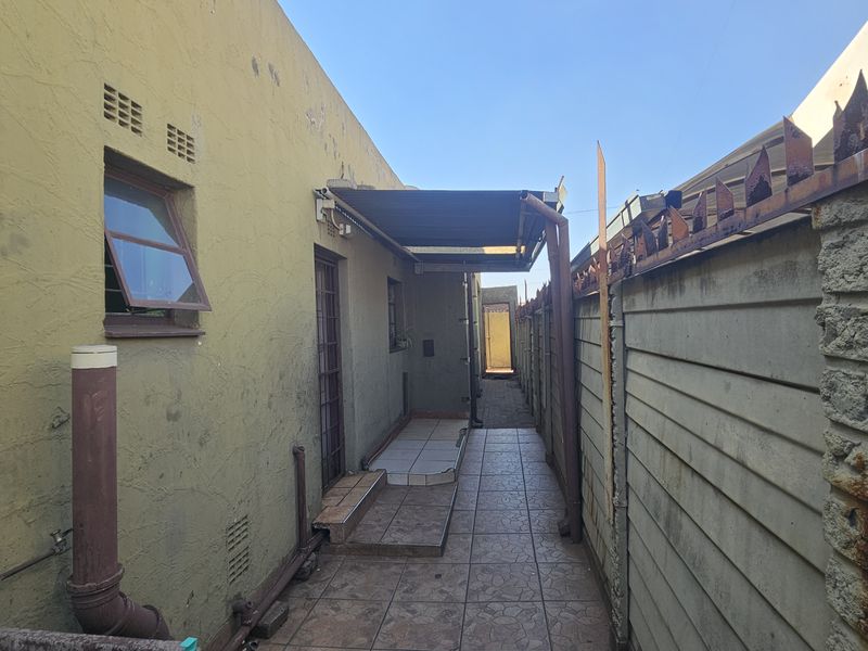 A narrow rear exterior walkway featuring tiled flooring, a covered section, and a precast concrete boundary wall with security spikes.