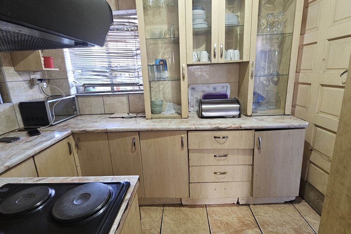 A dated kitchen featuring laminate cabinetry, built-in glass display units, tiled flooring, and an integrated electric hob with an extractor fan.