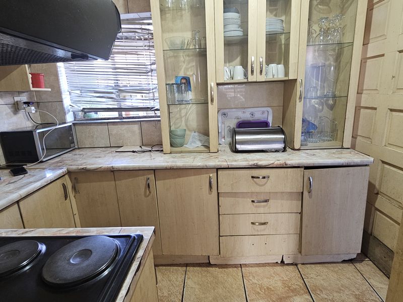 A dated kitchen featuring laminate cabinetry, built-in glass display units, tiled flooring, and an integrated electric hob with an extractor fan.