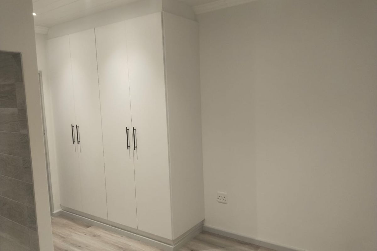 A clean, modern bedroom space featuring neutral walls, light laminate flooring, and ample built-in cupboard storage with contemporary hardware.