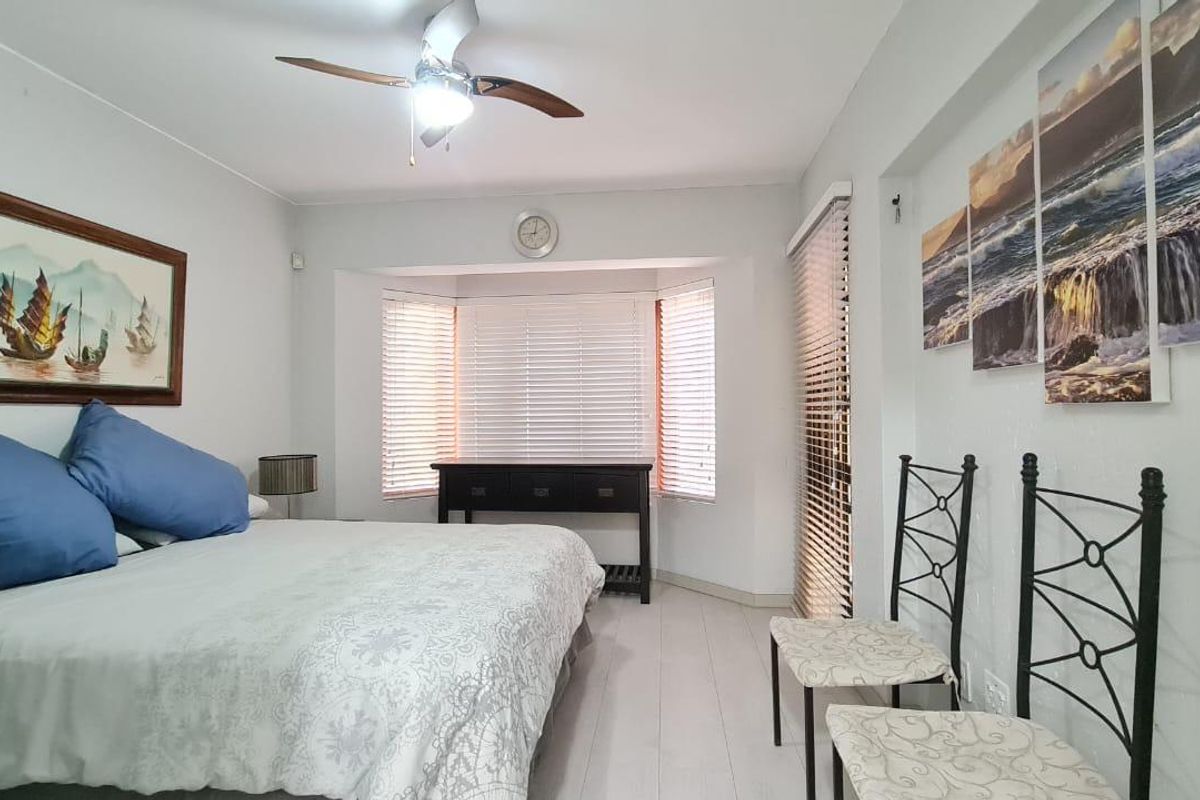 A well-lit bedroom featuring laminate flooring, a ceiling fan, and large windows with venetian blinds, providing a bright and comfortable living space.