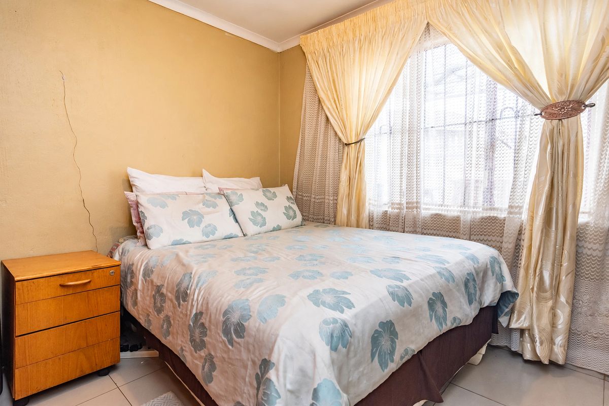A bedroom featuring tiled flooring and a window with curtain rails. The room shows signs of wear including a visible crack on the wall.
