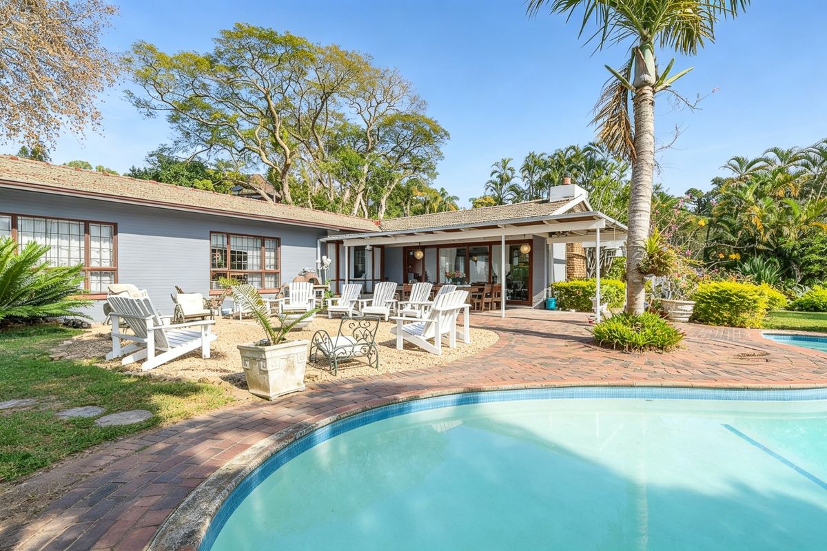 Spacious rear garden featuring a swimming pool, brick-paved patio, and a covered outdoor entertainment area set against a mature landscaped backdrop.
