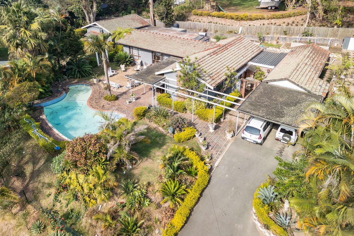 Aerial view of a residential property featuring a swimming pool, paved driveway, carport, and established garden surroundings.
