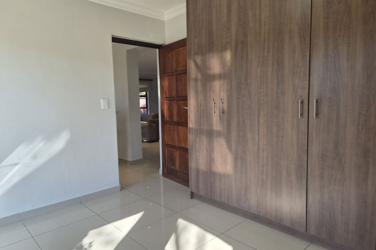 A bright, empty bedroom featuring modern wood-finish built-in cupboards, neutral tiled flooring, and a solid wood door leading to an adjacent living space.