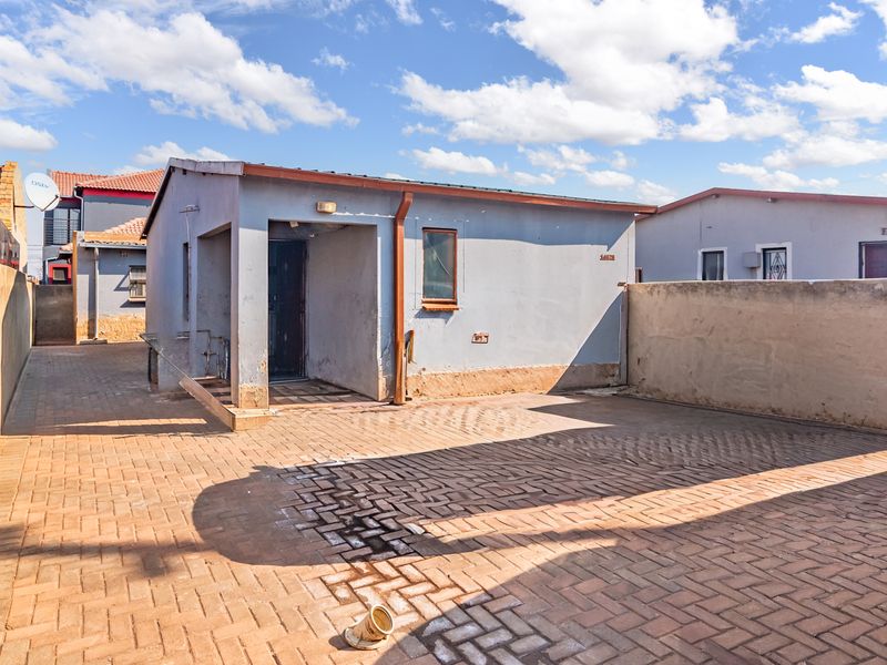 Rear exterior view of the property featuring a paved courtyard area, boundary wall, and basic building facade with visible maintenance requirements.