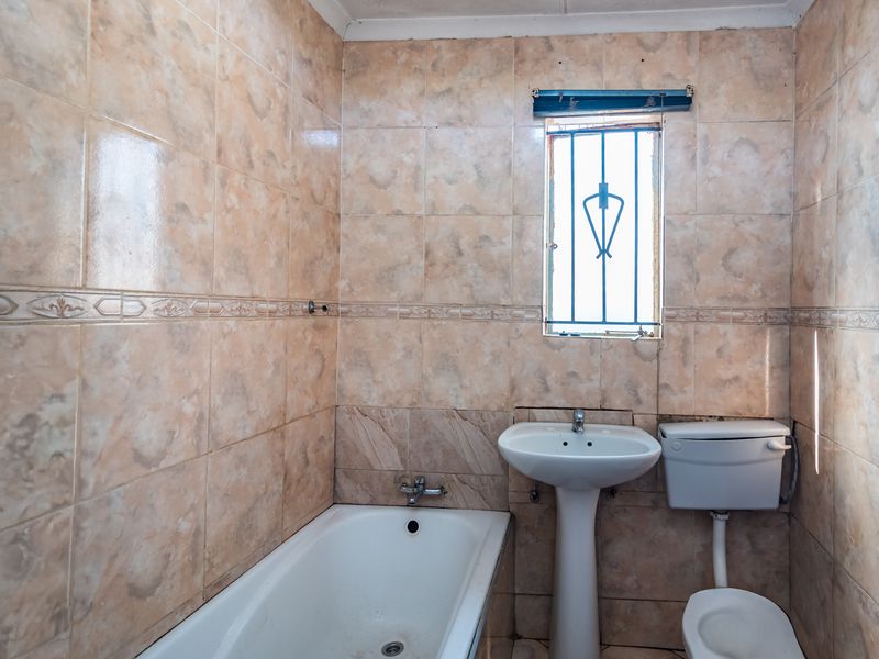 A dated bathroom featuring a pedestal basin, toilet, and bathtub with full-height wall tiling and a window with security bars.