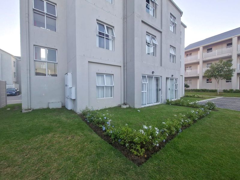 Rear view of a multi-story apartment building featuring a well-maintained lawn, landscaped garden beds, and sliding glass door access.